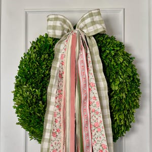 May include: A round, green boxwood wreath with a large bow made of green and white checkered fabric. The bow has several long, decorative ribbons in pink, cream, and green with floral and striped patterns. The wreath is hanging on a white door.