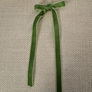 May include: A green velvet ribbon tied in a bow, with long, dangling tails. The ribbon is attached to a small, gold-colored ring. The bow and tails are a rich, solid green color.