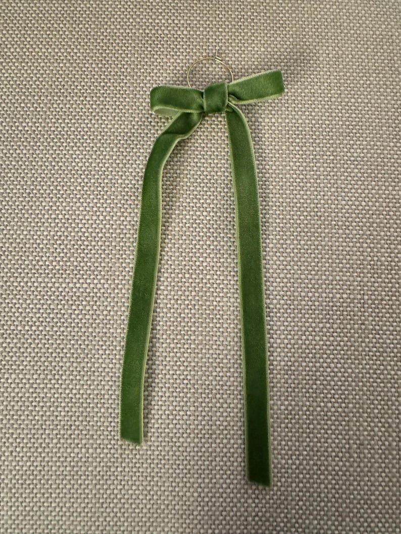 May include: A green velvet ribbon tied in a bow, with long, dangling tails. The ribbon is attached to a small, gold-colored ring. The bow and tails are a rich, solid green color.