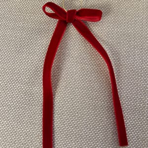 May include: A red velvet ribbon with a gold ring at the top. The ribbon is tied in a bow with two long, slender tails. The bow and tails are a deep, rich red color.