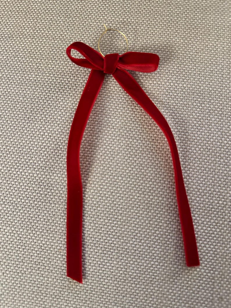 May include: A red velvet ribbon with a gold ring at the top. The ribbon is tied in a bow with two long, slender tails. The bow and tails are a deep, rich red color.