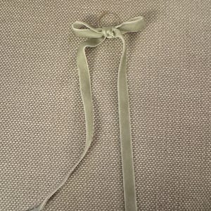 May include: A light green velvet ribbon tied in a bow with long, flowing tails. The ribbon is attached to a small, silver-colored ring. The bow and tails are centered against a textured, neutral-toned background.