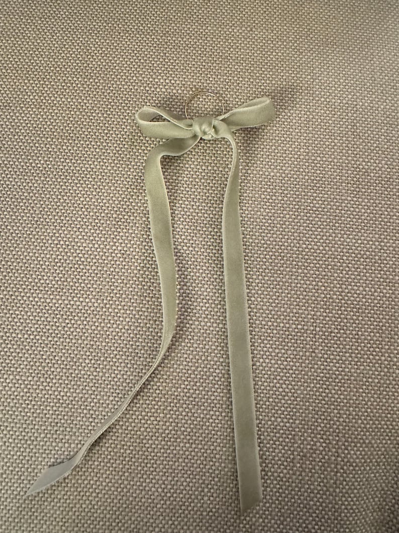 May include: A light green velvet ribbon tied in a bow with long, flowing tails. The ribbon is attached to a small, silver-colored ring. The bow and tails are centered against a textured, neutral-toned background.