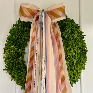 May include: A green boxwood wreath with a large bow made of cream and plaid ribbons in shades of orange, pink, and brown. Several ribbons in various colors and textures cascade down from the bow, including lace and solid-colored ribbons.