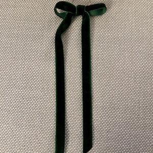 May include: A dark green velvet ribbon bow with long, flowing tails. The bow is attached to a small gold-colored ring. The ribbon has a soft, luxurious texture and is likely used for decoration or embellishment.