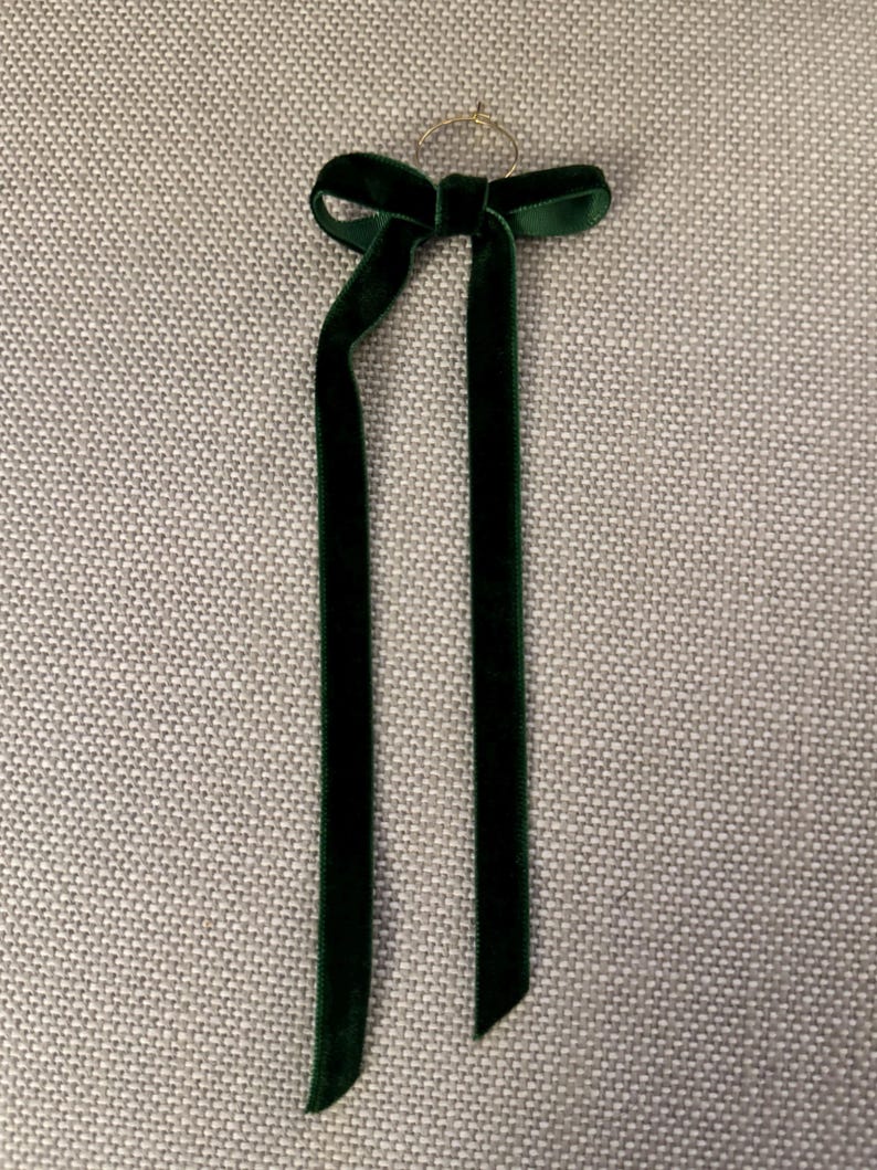 May include: A dark green velvet ribbon bow with long, flowing tails. The bow is attached to a small gold-colored ring. The ribbon has a soft, luxurious texture and is likely used for decoration or embellishment.