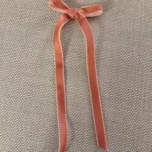 May include: A light pink velvet ribbon with a bow and long, flowing tails. The ribbon is attached to a small gold-colored ring. The item is likely used for hair accessories or gift wrapping.
