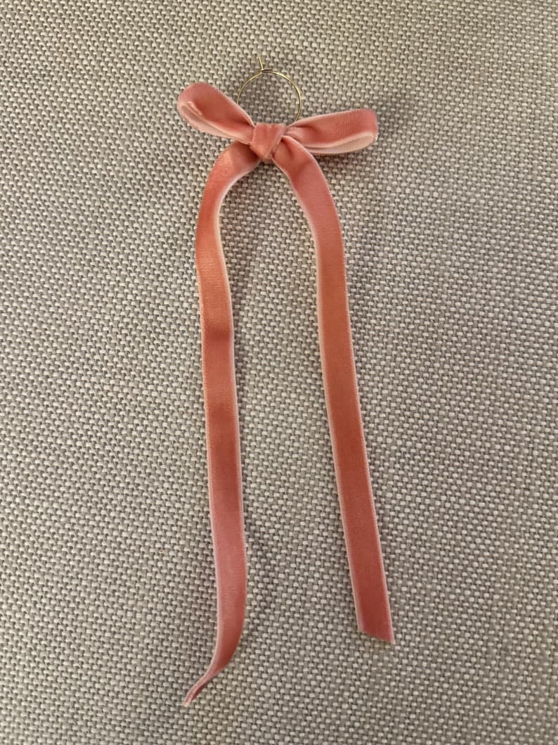 May include: A light pink velvet ribbon with a bow and long, flowing tails. The ribbon is attached to a small gold-colored ring. The item is likely used for hair accessories or gift wrapping.