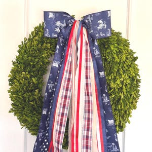 May include: A round, green boxwood wreath adorned with a large navy blue bow featuring a white toile pattern. Red, white, and blue ribbons cascade down, creating a patriotic and decorative accent for a door or wall.
