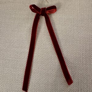 May include: A burgundy velvet bow with long, dangling streamers. The bow is attached to a small silver ring. The fabric has a soft, luxurious texture. The bow is likely used for decoration or as an accessory.