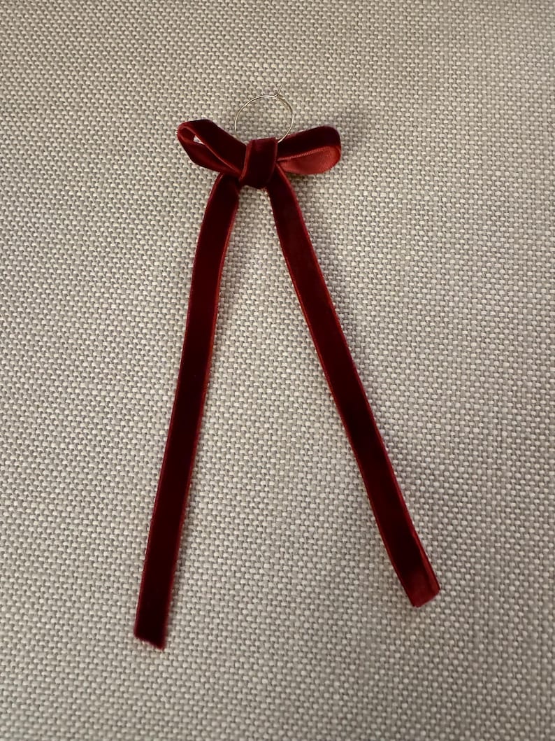 May include: A burgundy velvet bow with long, dangling streamers. The bow is attached to a small silver ring. The fabric has a soft, luxurious texture. The bow is likely used for decoration or as an accessory.