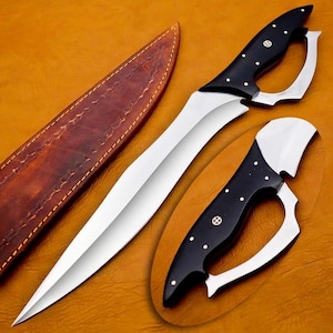May include: A handmade knife with a polished steel blade and a black handle with white dot accents. The knife has a unique curved design and is accompanied by a brown leather sheath. The overall design is sleek and functional.