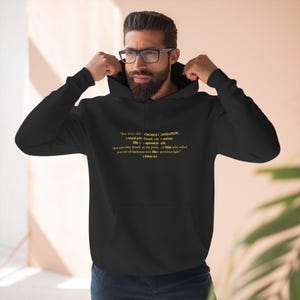 Peut inclure: Sweat à capuche noir avec cordon de serrage et poche avant. Le sweat à capuche présente un texte doré qui se lit "Mais VOUS ÊTES LA GÉNÉRATION CHOISIE..." de 1 Pierre 2:9. Le sweat à capuche est porté par une personne portant des lunettes.