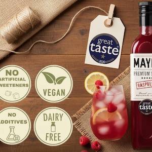 May include: A bottle of Mayer Premium Raspberry Syrup next to a glass of raspberry drink with ice and lemon slices. The image also shows a tag with the words "great taste 2020" and labels indicating the product is vegan, dairy-free, and has no artificial sweeteners or additives.