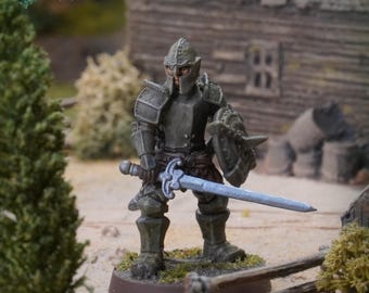 May include: A miniature figurine of a warrior clad in dark grey plate armor and a matching helmet. The figure holds a silver-toned sword in the right hand and a round shield in the left, standing on a circular base with textured ground details.