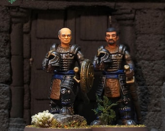 May include: Two hand-painted miniature figurines of warriors in dark metallic armor with blue sashes, each holding a helmet. The figures stand on circular bases with small tufts of artificial grass and foliage. The text IVYROCK is visible.