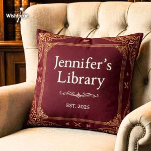 May include: A burgundy cushion with gold detailing and the text "Jennifer's Library" in white. The cushion has a decorative border and the text "EST. 2025". The cushion is on a beige armchair.