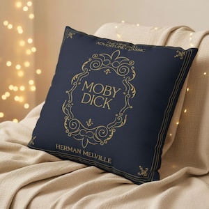 May include: A navy blue cushion with the title "MOBY DICK" in gold script within an ornate frame. The author's name, Herman Melville, is printed below. The cushion is a decorative accent for a home.