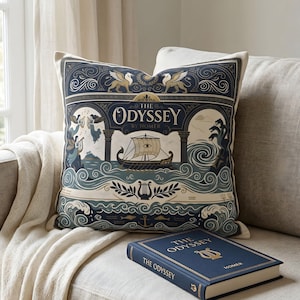 Puede incluir: Cojín decorativo con un diseño azul marino y crema que representa un barco, olas y el texto "The Odyssey by Homer". También se ve un libro a juego. El cojín está sobre un sofá beige.