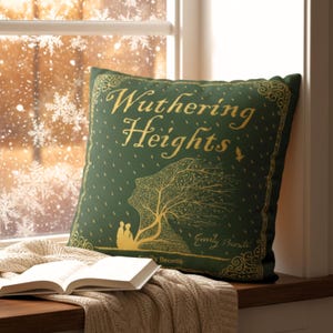 Puede incluir: Un cojín verde con letras y diseño dorados, con el título "Wuthering Heights" y una ilustración de un árbol y figuras. El cojín está colocado en un alféizar con un libro y una manta.