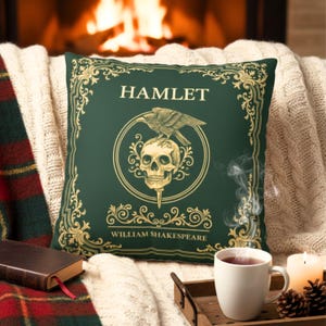 Può includere: Un cuscino quadrato decorativo con sfondo verde scuro e dettagli dorati. Il cuscino presenta la parola "HAMLET", un teschio con un corvo e le parole "WILLIAM SHAKESPEARE". Il cuscino è su una coperta color crema.