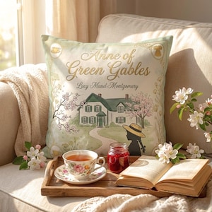 May include: A cushion featuring the text "Anne of Green Gables" and a watercolour illustration of a house and a woman in a hat. A teacup, jam jar, and open book are on a wooden tray.