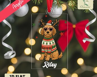 May include: A clear acrylic Christmas ornament with a cartoon dog wearing a festive sweater, heart-shaped sunglasses, and a Santa hat with antlers. The dog's name, "Riley," is printed below. The ornament hangs from a red ribbon.