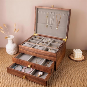 May include: A wooden jewelry box with multiple compartments and drawers. The box is open, revealing various necklaces, rings, bracelets, and a watch. The interior is lined with gray fabric. The box is made of dark wood and has gold-colored hardware.