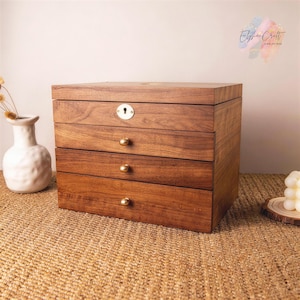 Könnte beinhalten: Eine rechteckige Schmuckschatulle aus Holz mit einer satten braunen Maserung, ausgestattet mit einem Deckel mit goldenem Schlüsselloch, drei horizontalen Schubladen mit kleinen goldenen Knöpfen und einer glatten Oberfläche.