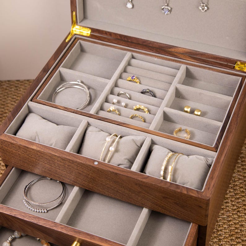 May include: A wooden jewelry box with multiple compartments and drawers. The interior is lined with gray fabric and holds various pieces of jewelry, including rings, bracelets, and earrings. The box is made of dark wood and has a hinged lid.