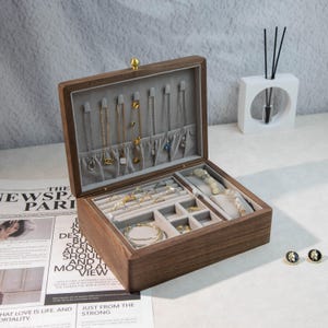May include: A wooden jewelry box with a grey velvet interior, showcasing necklaces, rings, bracelets, and earrings. The box is open, revealing various compartments for storage. A gold-coloured clasp secures the lid.