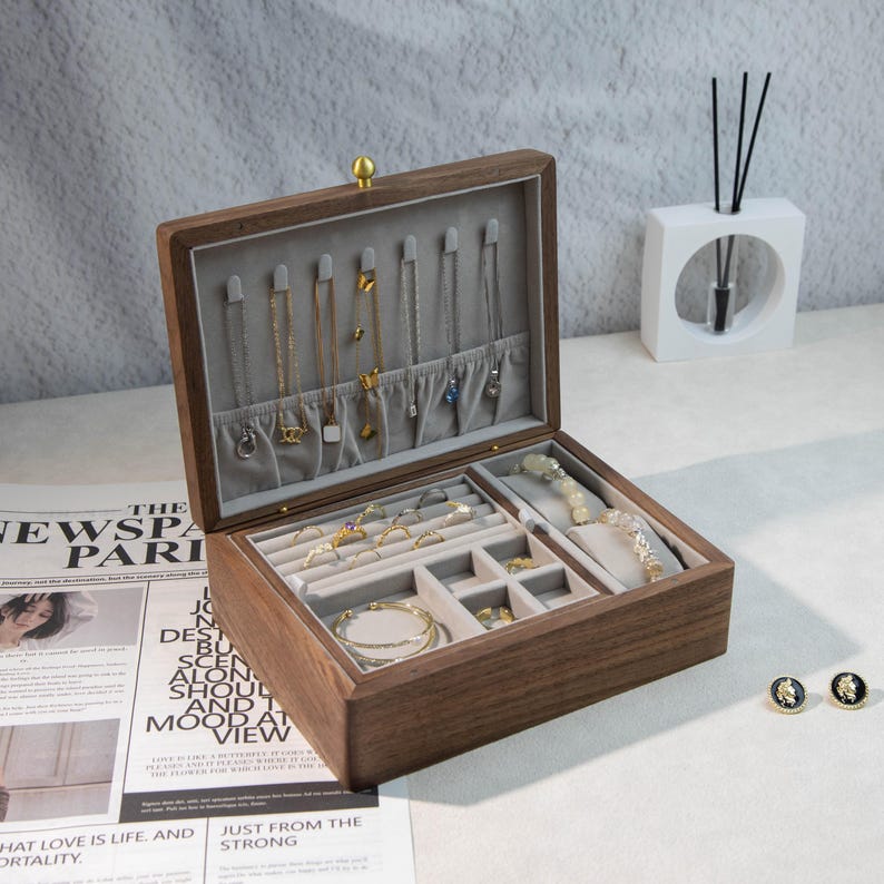 May include: A wooden jewelry box with a grey velvet interior, showcasing necklaces, rings, bracelets, and earrings. The box is open, revealing various compartments for storage. A gold-coloured clasp secures the lid.