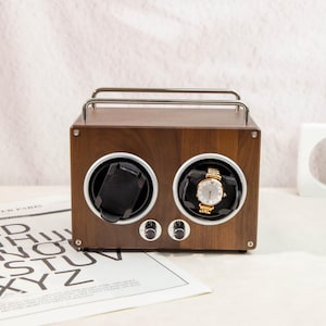 May include: A dark brown wooden watch winder with two compartments, each designed to hold a watch. The winder features silver metal accents and two control knobs. One compartment holds a gold watch with a white face.