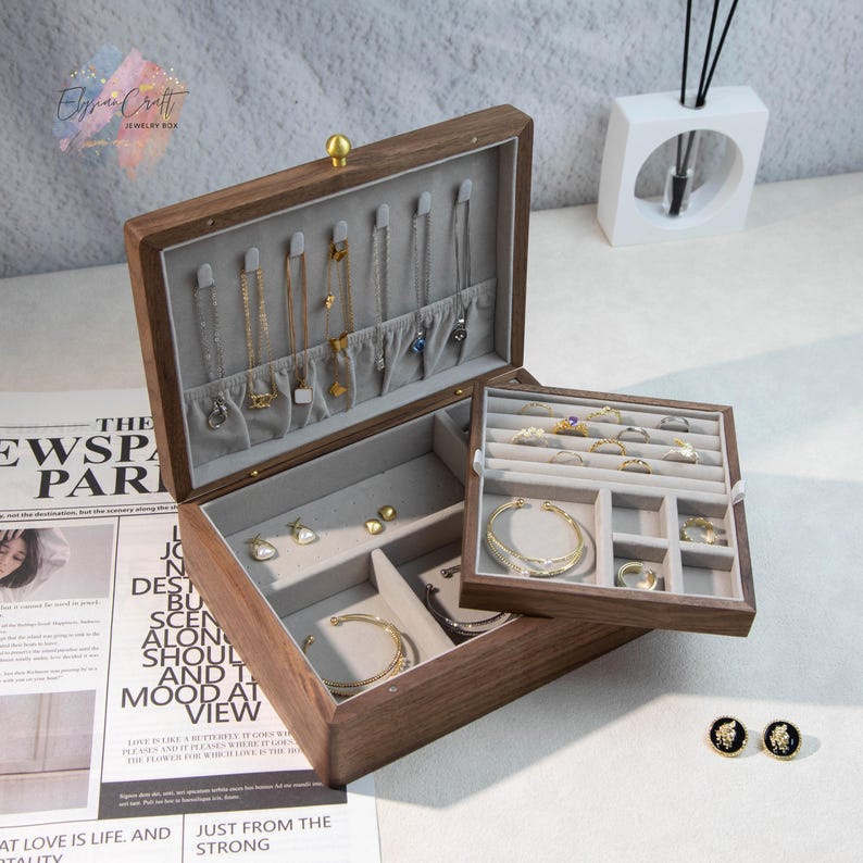 May include: A dark wooden jewelry box, open to display various jewellery pieces. The box has a hinged lid and is lined with grey fabric. Necklaces, bracelets, rings, and earrings are visible. A smaller tray is on top.
