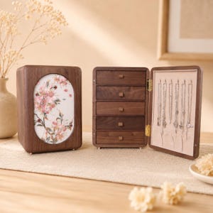 May include: A dark wood jewellery box with a closed side featuring an oval panel with pink floral artwork and an open side revealing five stacked drawers with small square pulls and a side panel with six silver hooks for hanging necklaces.