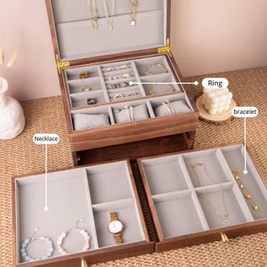 May include: A wooden jewelry box with multiple compartments, including sections for rings, bracelets, and necklaces. The box is open, revealing various pieces of jewelry. The interior is lined with gray fabric, and the box has a warm, natural wood finish.