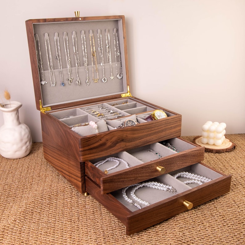 May include: A wooden jewelry box with multiple compartments and drawers. The box is open, revealing necklaces, rings, bracelets, and other jewelry. The box is made of dark wood and has a clear glass top. The drawers are lined with a soft, gray fabric.