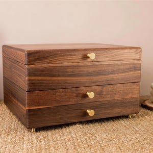 May include: A wooden jewelry box with three drawers and a top lid. The box is made of dark brown wood with a visible grain pattern. Each drawer has a small, round, gold-colored knob. The box sits on small gold feet.
