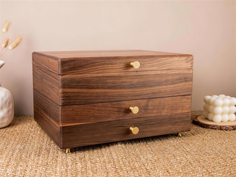 May include: A wooden jewelry box with three drawers and a top lid. The box is made of dark brown wood with a visible grain pattern. Each drawer has a small, round, gold-colored knob. The box sits on small gold feet.