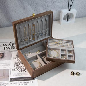 May include: A wooden jewelry box, open to show compartments lined with grey fabric, holding necklaces, bracelets, rings, and earrings. The box is made of dark wood and has a gold-coloured clasp. The box is open, revealing its contents.