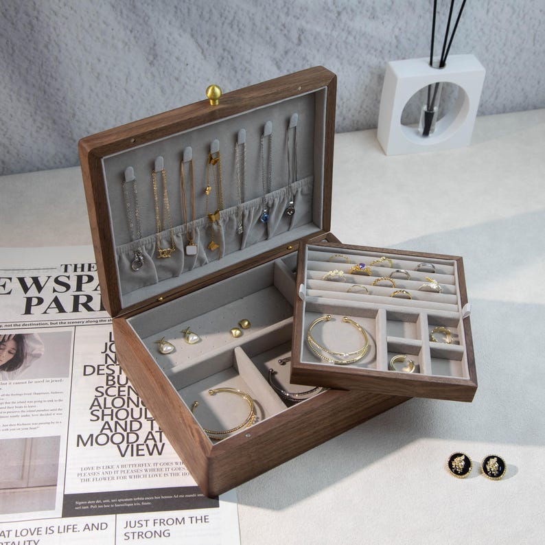 May include: A wooden jewelry box, open to show compartments lined with grey fabric, holding necklaces, bracelets, rings, and earrings. The box is made of dark wood and has a gold-coloured clasp. The box is open, revealing its contents.