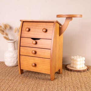 May include: A light brown wooden chest with four drawers, each featuring a single round knob. The top drawer has two knobs, while the lower three have one each. A curved wooden arm extends from the right side, supporting a small circular shelf.