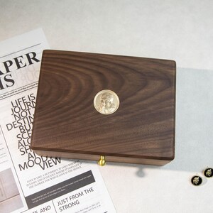 May include: A dark brown wooden box with a gold-coloured medallion featuring a religious figure. The box has a brass-coloured knob and is placed on a white surface with newspaper clippings. The medallion is circular.