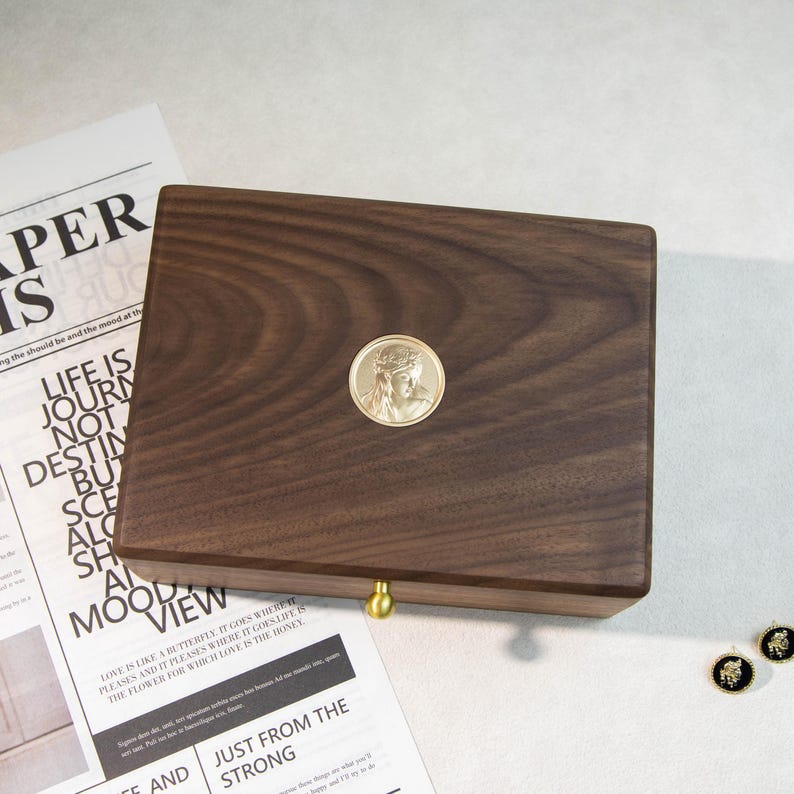 May include: A dark brown wooden box with a gold-coloured medallion featuring a religious figure. The box has a brass-coloured knob and is placed on a white surface with newspaper clippings. The medallion is circular.