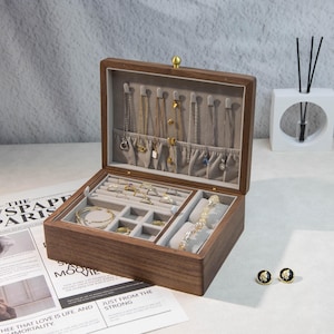 May include: A wooden jewellery box, open to reveal various compartments lined with grey fabric. The box contains necklaces, rings, bracelets, and earrings. The box is made of dark wood and has a gold clasp.