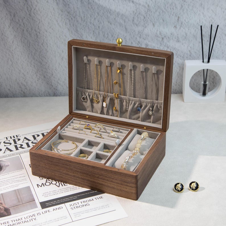 May include: A wooden jewellery box, open to reveal various compartments lined with grey fabric. The box contains necklaces, rings, bracelets, and earrings. The box is made of dark wood and has a gold clasp.