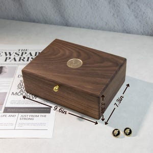 May include: A dark brown wooden jewelry box with a gold knob and emblem, measuring 24.4 cm long, 18.5 cm wide, and 8.4 cm high. The box is rectangular. Two black and gold earrings are next to the box. The text "THE NEWSPAPER PARIS" is visible.