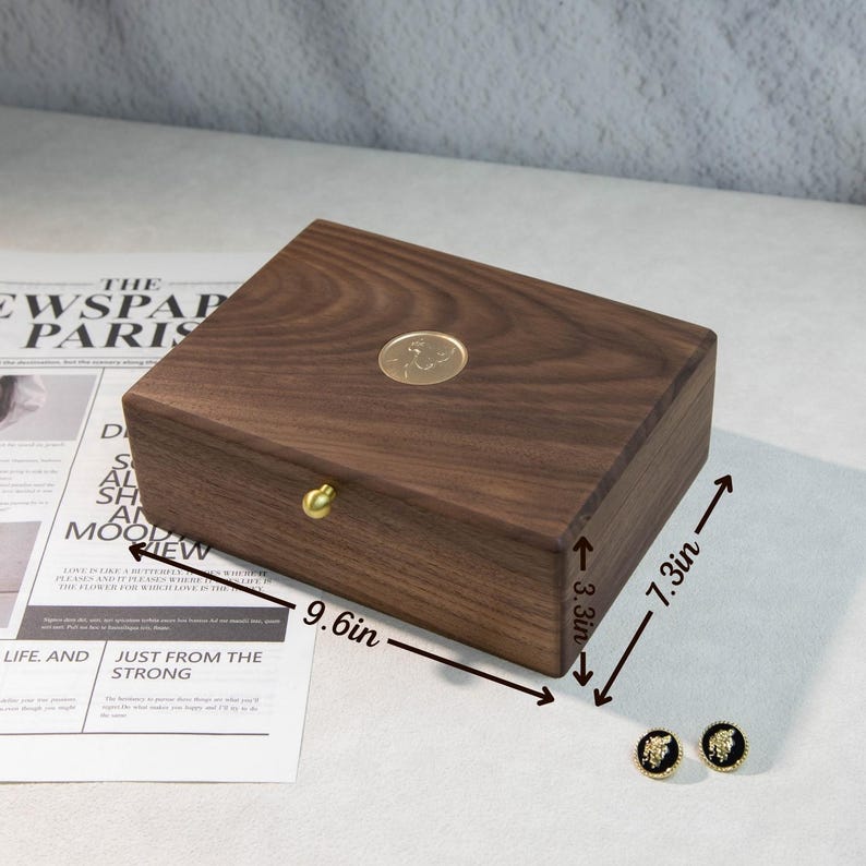 May include: A dark brown wooden jewelry box with a gold knob and emblem, measuring 24.4 cm long, 18.5 cm wide, and 8.4 cm high. The box is rectangular. Two black and gold earrings are next to the box. The text "THE NEWSPAPER PARIS" is visible.