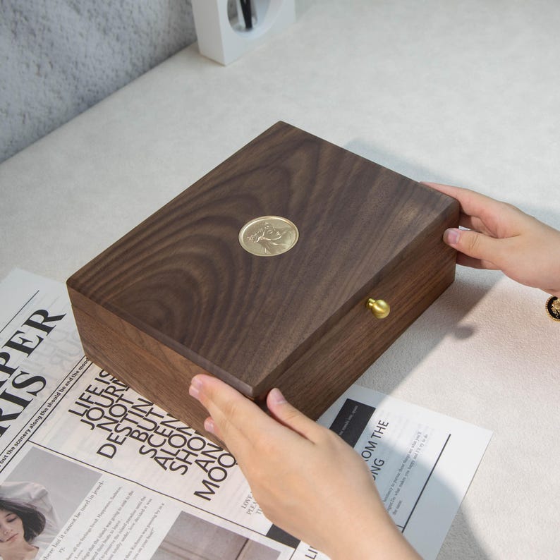 May include: A dark brown wooden box with a brass knob and a circular brass emblem on the lid. The rectangular box has a smooth, polished finish. The box is being held by two hands.