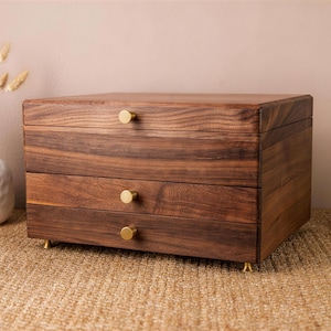 May include: A three-drawer wooden jewellery box with a rich brown finish. The box has gold-coloured knobs and feet. The wood grain is visible. The box is sitting on a woven surface.